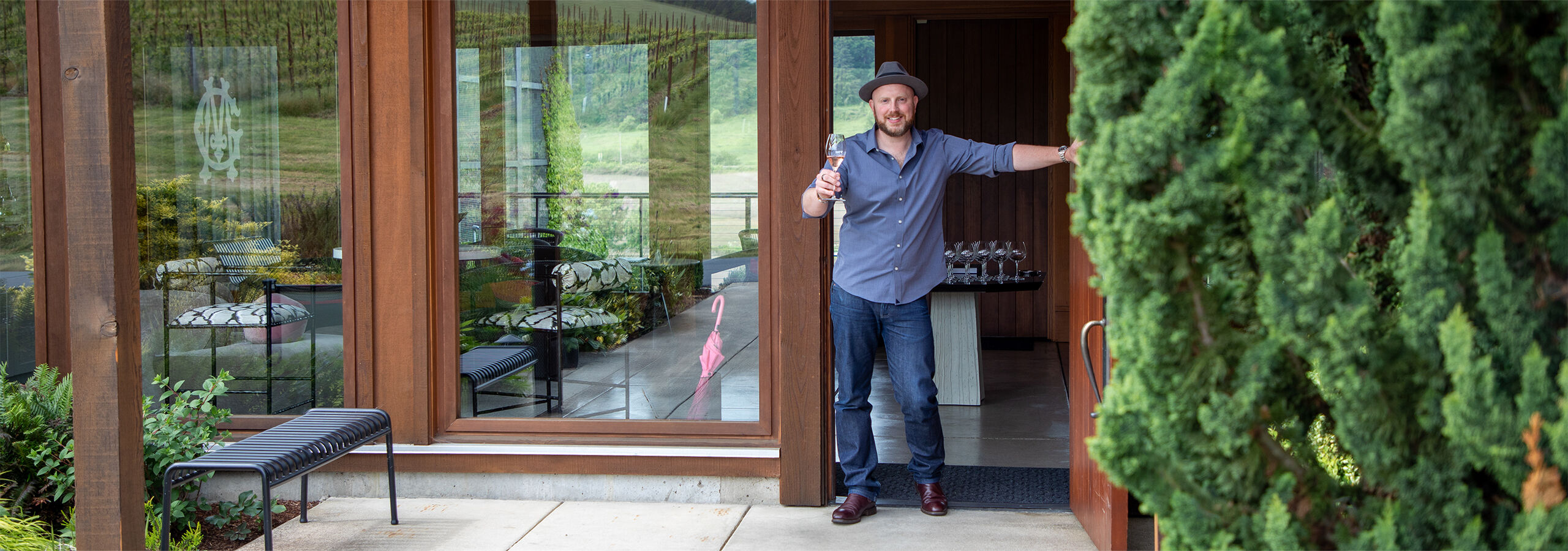 Estate Host with hat holding open the door of the Gran Moraine Tasting room with a glass of rosé wine outstretched as a greeting.